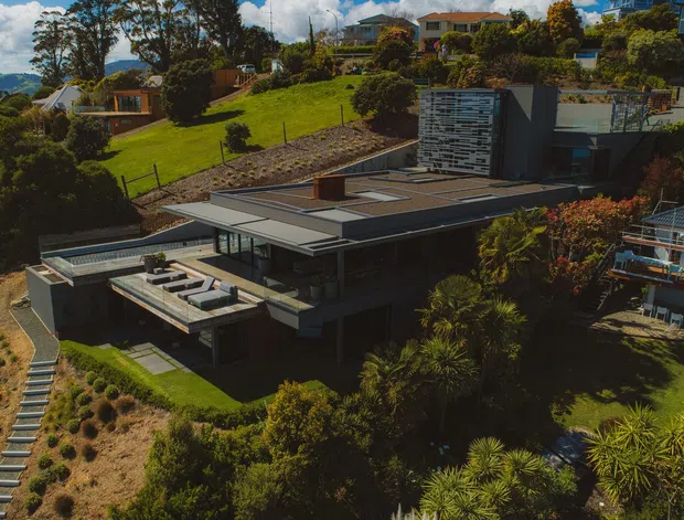 Aerial view of a contemporary multi-level house integrated into a steep, landscaped coastal or hillside site. The home has broad flat roof planes, extensive glazing, dark horizontal cladding, terraces, and a rooftop or upper-level courtyard. Dense planting surrounds the property, with neighbouring homes and a blue body of water visible in the distance.