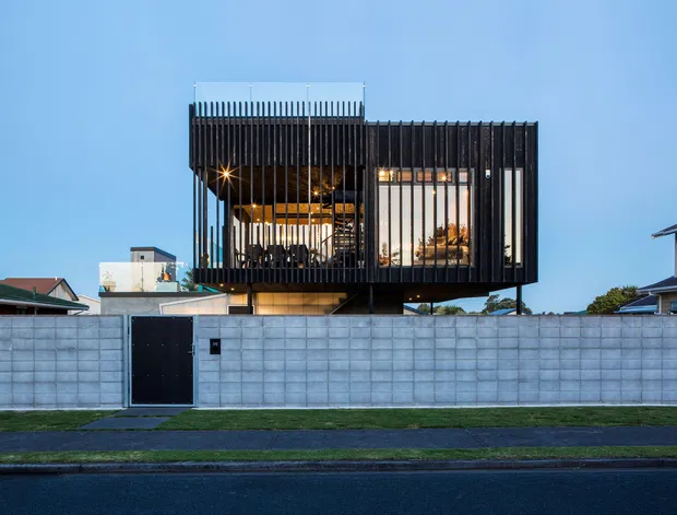 Street-facing view of a contemporary elevated two-storey residence behind a pale concrete block boundary wall. The upper volume is enclosed by a dramatic black vertical louvred screen, with warm interior lighting and glazing visible behind it. A flat roof and cantilevered form create a strong modern facade, with a dark pedestrian gate, paved footpath, lawn verge, and neighbouring suburban homes in the foreground and background.