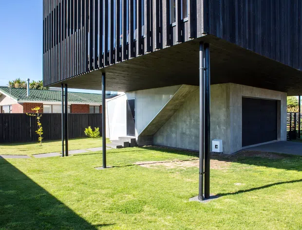 Exterior view of a contemporary elevated two-storey residence. A dark upper volume with closely spaced vertical slats projects over a concrete ground-level garage and entry zone, supported by slender dark posts. An external stair rises to the upper level, with a manicured lawn, dark fencing, and neighbouring suburban homes visible around it.