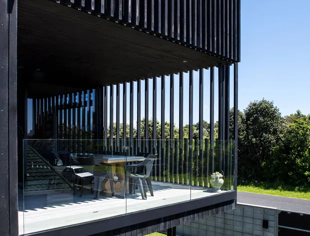 Exterior architectural photograph of a contemporary multi-level home with a dramatic cantilevered upper terrace. The structure features dark vertical timber battens, slender black supports, extensive clear glass balustrading, and an open outdoor dining area overlooking a landscaped lawn. Strong geometric lines, deep shadows, and a clear blue sky create a modern, sculptural appearance.