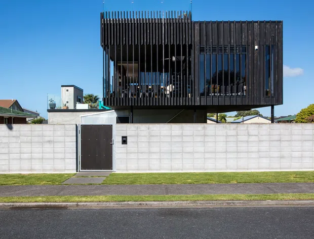 Street-facing view of a contemporary multi-level residence with a dark upper volume elevated above a pale concrete-block boundary wall. The upper facade combines extensive glazing with closely spaced vertical timber-like screening, creating a private, sculptural frontage. A simple pedestrian gate, narrow lawn, footpath, and residential surroundings are visible.