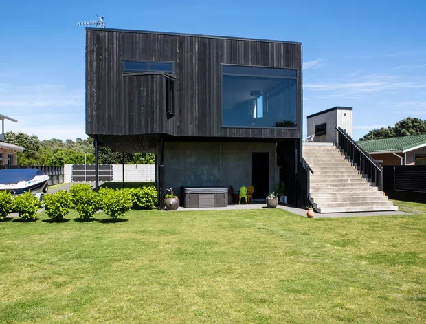 A modern two-storey residential extension or detached structure with a dark vertical timber-clad upper level, large glazed windows, and an exposed concrete lower level. An external concrete staircase leads to the upper floor, while the ground level opens onto a broad lawn with a covered outdoor area. The setting is suburban, with neighbouring homes and fences visible.