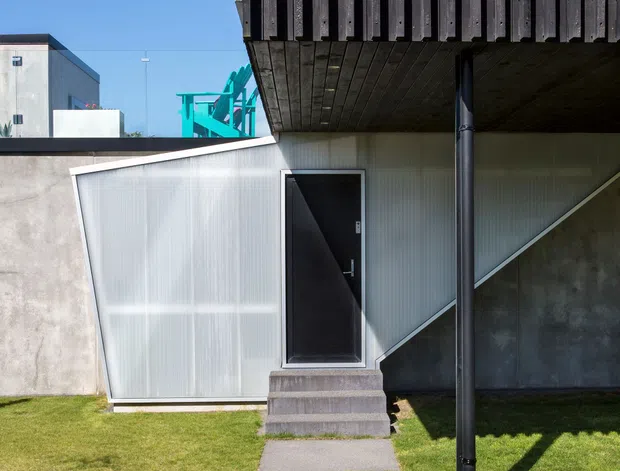 Exterior view of a compact contemporary building entrance, featuring a black door reached by concrete steps and a simple paved path through lawn. The facade combines translucent corrugated cladding with exposed concrete, while a projecting upper balcony has dark timber or metal vertical slats and a timber-lined underside. Strong geometric forms, an angled roofline, and an external black support post create a modern architectural composition under clear daylight.