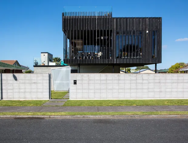 Street-facing view of a contemporary multi-level residence with a dark, box-like upper volume screened by closely spaced vertical fins and extensive glazing. The upper storey appears cantilevered over a recessed lower level, behind a pale concrete block boundary wall with a pedestrian gate. Neighbouring suburban homes and a paved street are visible.