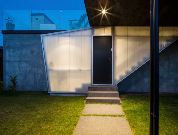 Twilight exterior view of a compact modern building entry. A dark metal door is centered within a translucent ribbed panel façade, with exposed concrete walls, a projecting dark timber-lined roof canopy, an external stair enclosure, and a simple concrete path across a manicured lawn. The composition emphasizes contemporary materials, geometric forms, and warm entry lighting against a blue evening sky.