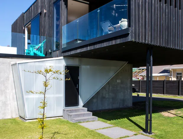 Exterior view of a contemporary multi-level residence with a strongly cantilevered upper floor, dark vertical timber screening, recessed balconies, and clear glass balustrades. The lower level combines concrete and corrugated metal surfaces, with a small landscaped lawn and stepping-stone path leading to the entrance. Bright daylight and a vivid blue sky emphasize the crisp modern facade.