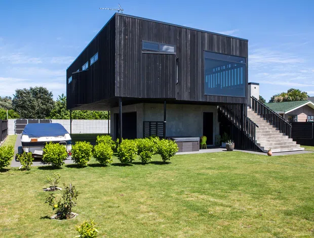 Exterior view of a contemporary two-storey residence with a strongly cantilevered upper volume clad in dark vertical timber boards. The lower level has a concrete-like finish, while an external stair leads to the upper floor. Large windows, a manicured lawn, hedges, boundary fencing, and a partially visible carport/driveway complete the suburban setting.