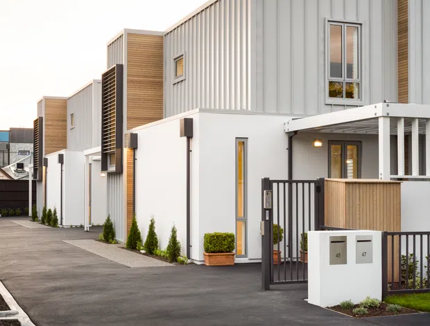 Contemporary multi-unit residential development viewed from a paved access lane. The buildings feature crisp white rendered walls, vertically ribbed metal cladding, warm timber-look vertical panels, projecting upper volumes, narrow vertical windows, and dark metal gates and railings. Landscaped planter strips and letterboxes define the foreground entry.