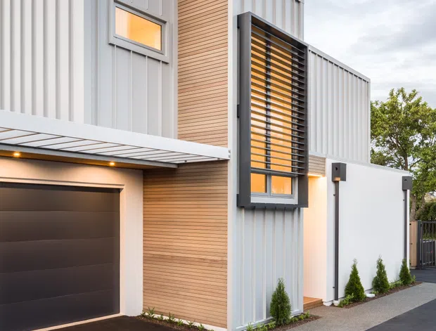 Contemporary multi-level residential facade viewed from the driveway, featuring pale vertical metal cladding, warm horizontal timber-look wall panels, projecting dark louvres over a tall window, a flat-roofed entry canopy, and an integrated dark garage door. Minimal planting and paved surfaces complete the clean urban exterior.
