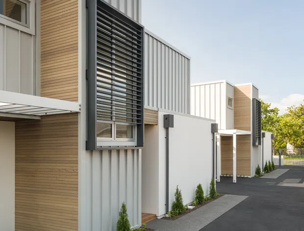 A contemporary multi-unit residential exterior with a linear, stepped form. The facades combine pale corrugated metal cladding, warm horizontal timber-look wall panels, white rendered sections, and dark aluminium louvred window screens. A paved asphalt driveway and narrow landscaped planting strips run alongside the buildings, with strong afternoon shadows emphasizing the architectural rhythm.