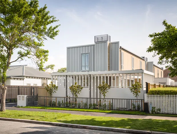 Street-facing view of a contemporary multi-level residential house with a grey vertical-panel upper storey, pale lower facade, extensive white-framed glazing and a covered veranda or screened outdoor walkway. The property sits on a landscaped suburban street with fencing and established trees.