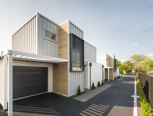 Bright exterior view of a contemporary multi-unit residential development with vertically ribbed light cladding, timber-look wall panels, projecting sunshade screens, carports, garages and a narrow paved access lane. The clean geometric forms and repeated townhouse-like units create a modern urban infill character.