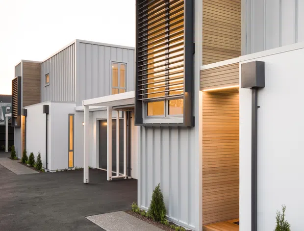 Exterior view of a row of compact modern multi-storey dwellings with crisp geometric forms. The façades combine pale grey vertical metal cladding, warm horizontal timber-look wall panels, white projecting elements, dark window louvres, and narrow glazed openings. A paved pedestrian path and dark asphalt driveway run along the buildings, with small planted shrubs at the entrances.