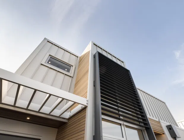 Low-angle exterior view of a contemporary multi-level building, featuring crisp white metal-clad volumes, vertical timber-look wall cladding, a prominent dark horizontal louvred screen, high windows, and a translucent covered entry or terrace canopy. The composition emphasizes angular rooflines and layered facade elements against a bright sky.