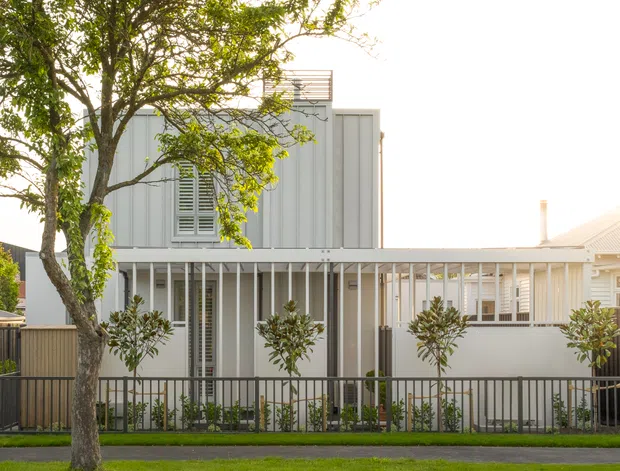 Street-facing view of a contemporary residential house with a light grey vertically clad upper storey, a long covered verandah with slim white posts and railings, shuttered windows, and a low front fence. Mature street trees and neatly planted shrubs frame the facade.