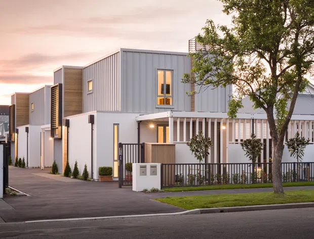 Street-level exterior view of a contemporary multi-unit residential development with attached townhouse-like dwellings. The buildings feature light grey vertical metal cladding, white rendered sections, timber-toned accents, narrow windows, covered entry porches, vertical screening, front fences and landscaped verge planting. The image is taken at dusk in a suburban streetscape.