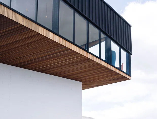 Exterior architectural detail of a contemporary building with a projecting upper volume, dark vertical metal cladding, extensive glazed windows, and a warm timber-lined soffit. A clean white masonry wall supports the cantilevered form, with modern exterior lighting and landscaped surroundings visible below.