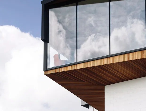 Close-up exterior architectural view of a contemporary multi-level building. A dramatic cantilevered upper volume features extensive floor-to-ceiling glazing in dark metal frames, a warm timber-lined soffit, and vertical timber cladding beneath the windows. White masonry walls and a curved metal-balustraded balcony are visible below, set against a bright sky.