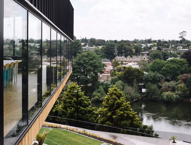 Exterior architectural view of a contemporary multi-level building overlooking a leafy urban waterway. The facade combines extensive floor-to-ceiling glazing with dark vertical cladding and warm timber-toned horizontal trim. A landscaped terrace and balustrade sit below, while the reflective glazing reveals a sparse interior with colourful vertical furnishings. Dense trees, residential buildings, and water form the surrounding setting.