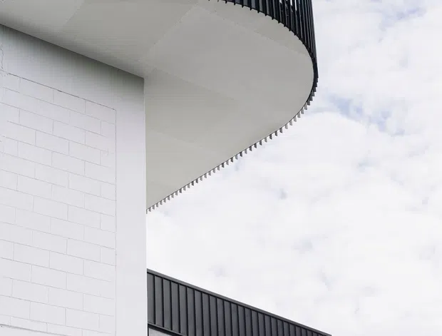 Close-up upward view of a contemporary building exterior featuring a dramatic curved balcony with slender black metal balustrades, white rendered and block-patterned walls, and a projecting glazed upper volume. Warm timber soffit lining contrasts with the monochrome facade, while the building is set against a bright cloudy sky.