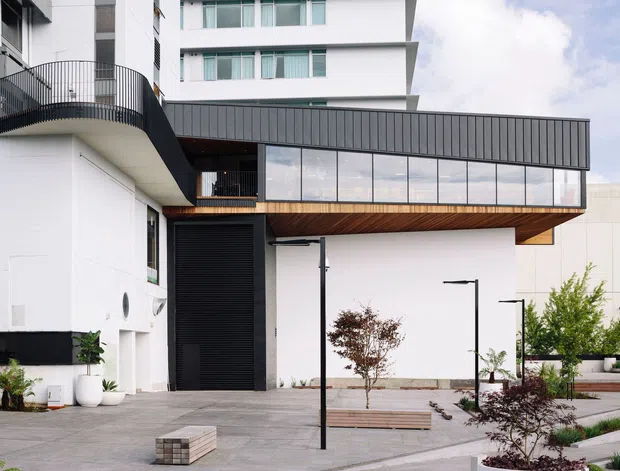 Exterior view of a contemporary multi-storey building with a prominent cantilevered upper volume, dark vertical metal cladding, extensive glazing, white rendered walls, timber-lined soffit, balconies, and a landscaped paved forecourt.