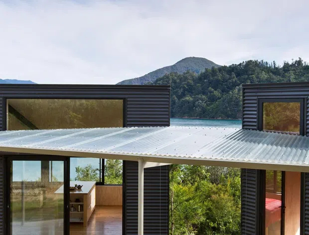 A modern single-storey house viewed from a broad timber deck, with dark horizontal cladding, extensive black-framed glazing, a translucent corrugated roof and open-plan indoor-outdoor connections. Through the glazing, a kitchen and a bedroom are visible. The house sits in a lush mountainous setting beside a bright blue body of water.