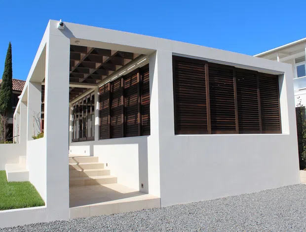 A modern white-rendered residential extension or pavilion viewed from the exterior, featuring a flat roof, deep covered entry, exposed timber ceiling beams, and large dark timber louvred window screens. Pale tiled steps lead into the structure, with gravel landscaping and neighbouring residential buildings visible around it.