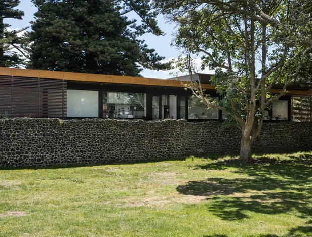 Single-storey low-profile modern residence viewed from the garden, with a long horizontal form, dark timber or metal louvred screening, extensive dark-framed glazing, and a warm-toned flat or very low-pitched roof edge. A distinctive dark rounded-stone masonry wall runs across the foreground, partially screening the house. Mature trees and lawn create a landscaped suburban setting.