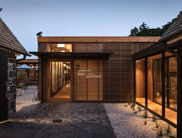 Twilight exterior view of a contemporary pavilion or small building complex framed by dark stone-clad structures. The central timber-clad pavilion has horizontal slatted screens, warm interior lighting, glazed doors and windows, and a low flat roof. Irregular dark stone paving leads through a courtyard, with pale gravel landscaping along the glazed right-hand wing.