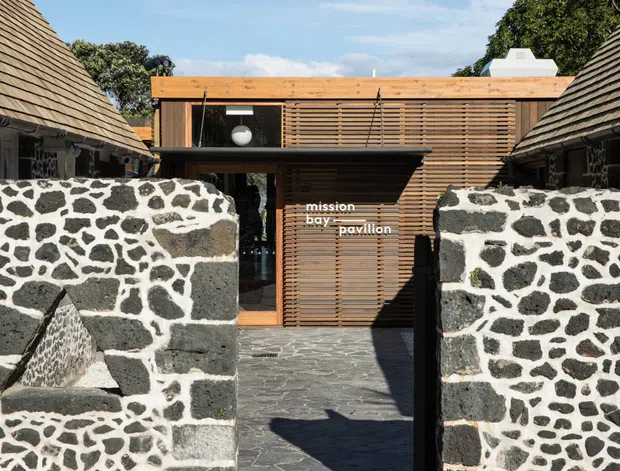 Exterior view of a small pavilion entrance framed by dark volcanic-stone masonry walls. The central building has warm timber cladding and horizontal timber screening, with a low projecting roof and glazed entry. Matching steeply pitched shingle-roof structures flank the passage, creating a courtyard-like approach.