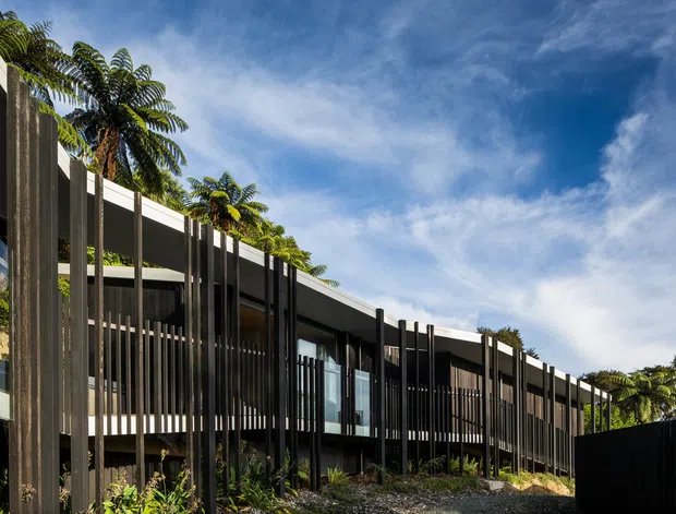 Exterior architectural view of a long, low-slung contemporary building with a gently curved flat roofline, extensive glazing, and repeated dark vertical screening elements. The structure is set among tropical planting and landscaped gravel, with a bright blue sky visible above.
