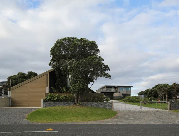 Street-level exterior view of a low-rise residential property in a suburban setting. The main building has a simple steeply pitched gable roof, warm timber-like horizontal cladding, and a dark recessed entrance or side wall. A large established tree, low stone retaining wall, gravel driveway, metal gate, lawn, and neighbouring homes are visible. The scene is overcast with a restrained, contemporary residential character.