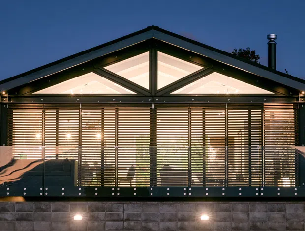 Night-time exterior view of a compact single-storey pavilion or house with a prominent gable roof, illuminated clerestory-like triangular glazing, extensive horizontal timber-look louvres or blinds across the front facade, dark structural framing, and decorative side panels. The building sits on a raised concrete block base with integrated exterior lights and is surrounded by vegetation.