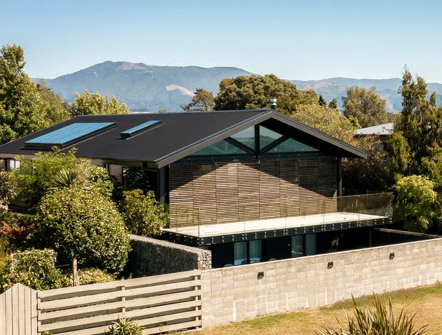 Exterior view of a contemporary single-storey residence with a broad dark gabled roof, multiple skylights, extensive glazing, timber screening and a pale masonry boundary wall. The home is set among established landscaping with mountain ranges visible in the background.