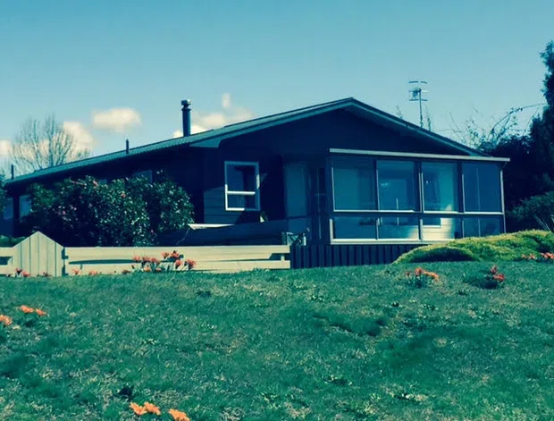 Single-storey dark-clad house with a simple pitched gable roof, enclosed glazed sunroom, broad lawn, and modest garden planting. The image has a slightly vintage, rural-suburban character.