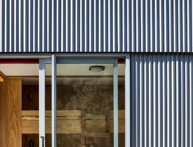 Close-up exterior view of a compact modern building clad in dark grey-blue vertical corrugated metal siding. A glazed sliding door opens to a warm timber-lined interior with built-in bunk-style seating or sleeping platforms, while a timber deck runs along the facade. The image emphasizes the ribbed metal envelope, large glass entry, and contrasting natural wood interior.