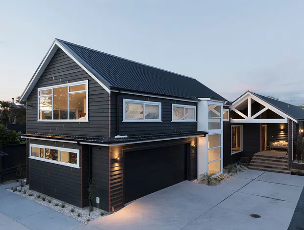 Exterior view of a contemporary dark-clad residential house with a steep gabled roof, attached double garage, concrete driveway, and a recessed illuminated entry beneath a prominent timber-trussed gable. The facade combines dark horizontal cladding with light trim and large glazed windows.
