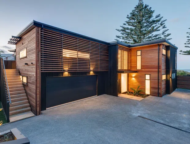Exterior view of a contemporary multi-level residence with dark timber cladding, extensive horizontal timber screening, and dark metal roof and garage elements. The stepped form includes an external stair, upper-level glazing behind louvers, illuminated recessed entry areas, and a broad concrete driveway or terrace. Mature conifer trees and a glimpse of the surrounding landscape frame the house.