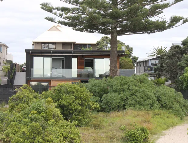 Exterior view of a contemporary two-storey residential home set within dense coastal-style landscaping. The dark upper-level facade combines broad glazing with warm timber cladding, while a pale pitched roof and mature Norfolk Island pine dominate the streetscape. Neighbouring homes and a sandy footpath are visible around the property.