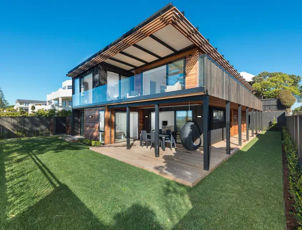 Modern two-storey residential home viewed from the landscaped backyard, featuring extensive floor-to-ceiling glazing, dark vertical cladding, timber detailing, a projecting upper-level terrace, and a covered ground-floor outdoor living area. The building has a strong geometric form with a pitched/sloping roof edge and a private fenced lawn.