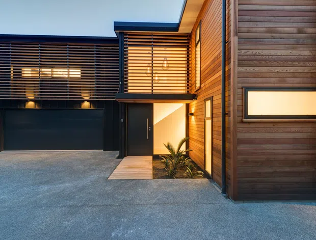 Contemporary multi-level residential facade viewed from the driveway at dusk. The exterior combines dark horizontal cladding and timber weatherboards, broad timber-framed windows with horizontal blinds, a dark garage door, recessed entry, warm exterior lighting, and a small planted entry bed.