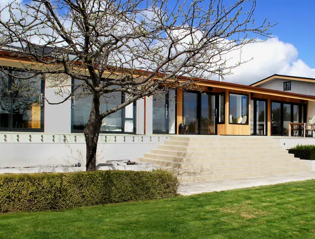 A low-slung modern single-storey residence viewed from the landscaped front lawn. The home has a mostly white rendered facade, broad floor-to-ceiling glazing, warm timber-framed windows and doors, a shallow pitched roof, and a raised terrace reached by wide pale stone steps. Trimmed hedges and a mature deciduous tree sit in front of the building under a bright blue sky.