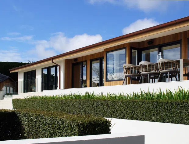 Single-storey contemporary residential home with a long low-pitched roof, warm timber-clad facade, extensive dark-framed glazing, and a raised outdoor dining terrace. Crisp white rendered walls and neatly clipped hedges create a clean, landscaped exterior composition under a bright blue sky.