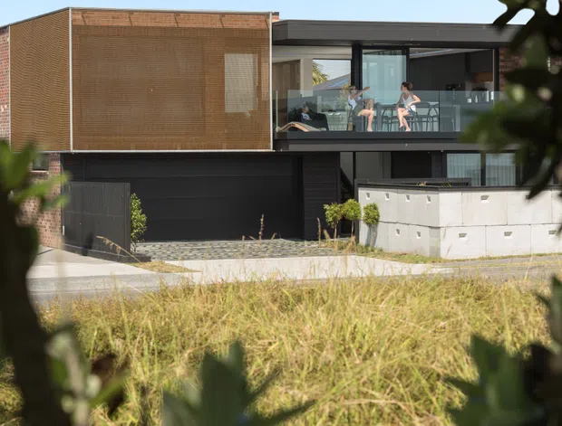 Exterior view of a contemporary multi-level residence framed by foreground foliage. The house features a flat roof, dark lower-level cladding, a large glazed upper balcony, a warm brown textured screen or cladding volume, a dark garage door, and a low pale concrete retaining wall. A sloping grassy site and paved driveway are visible in front.