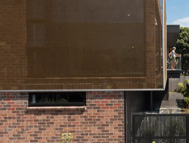 Exterior view of a contemporary urban residence featuring a red and brown brick lower facade, a large upper enclosure screened with fine bronze perforated metal mesh, dark metal framing, and a black vertical-slat gate. Low landscaping and a neighbouring balcony are visible at the edges.