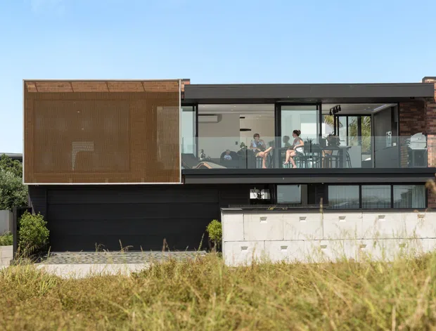 Exterior view of a contemporary multi-level residence with a flat roof, dark horizontal cladding, expansive glazing, a projecting upper-level balcony, and a prominent timber screen enclosing the left side. A pale masonry boundary wall and landscaped foreground sit in front of the house.