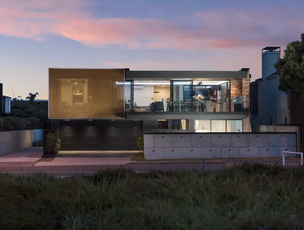 A contemporary multi-level residence viewed from the street at dusk. The house features a rectilinear composition, extensive glass walls, a warm timber-clad upper volume, dark garage doors, concrete retaining or boundary walls, and an open balcony or living area. Dense vegetation and neighbouring urban buildings frame the property.