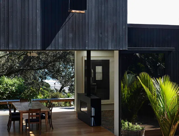 Exterior view of a contemporary dark-clad house with a projecting upper volume, large glazed openings, and an open timber deck. The outdoor living area includes a dining setting, lounge chairs, a small fire pit or low table, timber decking, gravel landscaping, and tropical planting. Strong sunlight creates dramatic shadows across the vertical cladding and deck.