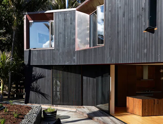 Exterior view of a compact two-storey home set among mature vegetation, with dark vertical timber cladding, angled roof planes, large windows, and a timber deck. The deck includes two lounge chairs and a small round table, while an open ground-level section reveals a warm timber-lined kitchen or living area. A planted raised garden edge and dark slatted screening contribute to the contemporary, natural setting.