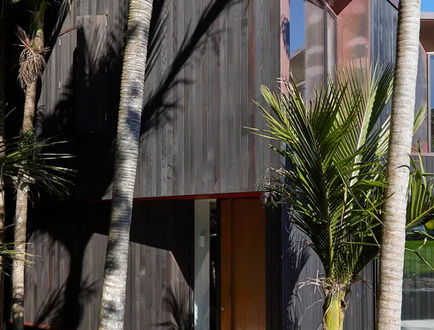 Exterior view of a compact contemporary multi-level building clad in dark vertical timber boards, with rust-toned metal roof and trim, large glazing, and a timber deck surrounded by palm and coastal vegetation. Strong geometric roof forms and deep shadows give the facade a modern architectural character.